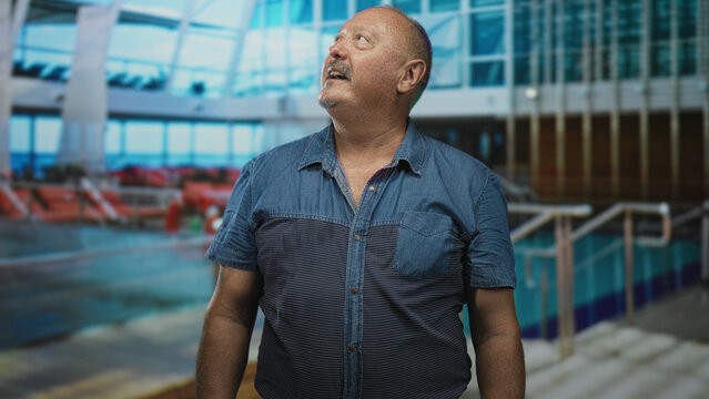 Man looking up with relaxed smile, denim shirt and mustache visible, standing by pool railing inside a building; calm reflection.