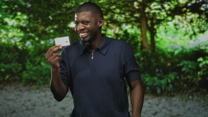 Man holding creditcard between fingers, smiling and presenting the chip card while wearing a navy polo shirt in a forest setting; confidence trust.