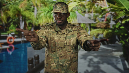 Soldier man in camouflage uniform stands with arms crossed gesture and steady gaze by a pool lifebuoy on a tropical resort backdrop in a studio  duty pride. © Krakenimages.com