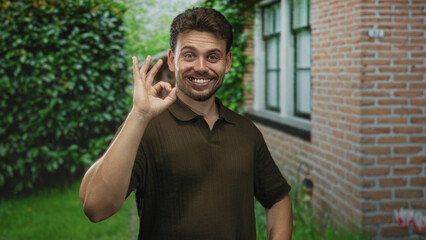 Man smiling with hands on hips by a building window at home entrance, facing camera; welcoming...