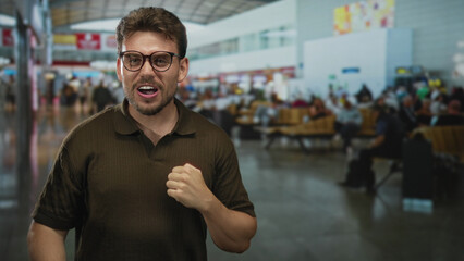 Obraz premium Man wearing glasses clenching fist and grimacing beside departure gates in a crowded airport terminal; frustration travel delay.