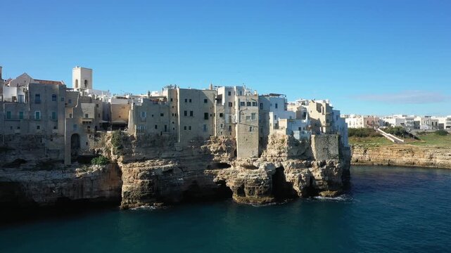 Historic stone buildings perched on dramatic cliffs above the Adriatic Sea in Polignano a Mare, Italy under a clear blue sky.