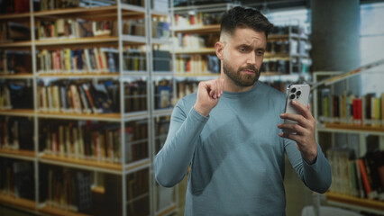 Man holding smartphone, checking the screen with a subtle wave gesture among bookshelves inside a building  pensive doubt. © Krakenimages.com
