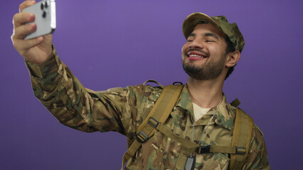 Hispanic soldier in camouflage uniform wearing dog tags holds smartphone at arm in purple studio; pride duty.