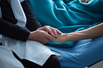 Close up of Caucasian female doctor holding patient hand with intravenous line attached in hospital bed, offering comfort and reassurance during inpatient treatment in modern healthcare setting.