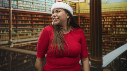 Fototapeta premium Woman in santa hat smiles and raises fists in a library building while turning her head and showing bare forearms; holiday joy.