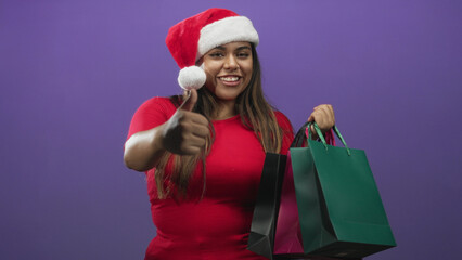 Fototapeta premium Woman holding shopping bags and wearing a santa hat in studio with alternating neutral and smiling poses including a thumbs up gesture; holiday cheer.