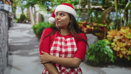 Fototapeta premium Woman wearing santa hat and red gingham apron, arms crossed holding elbow while gazing sideways in studio holiday set; playful holiday mood.