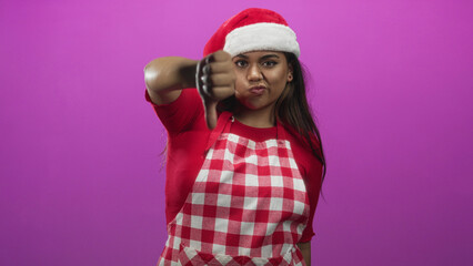 Fototapeta premium Woman wearing santa hat and red gingham apron gives thumbs down to camera in studio with magenta backdrop; holiday disapproval.