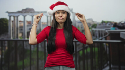 Fototapeta premium Woman wearing santa hat and red shirt raising hands by head on balcony of an old building; holiday frustration.