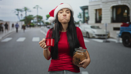 Fototapeta premium Woman in santa hat holding red mug and cookie jar on a street crosswalk, smiling and offering cookies; holiday joy.
