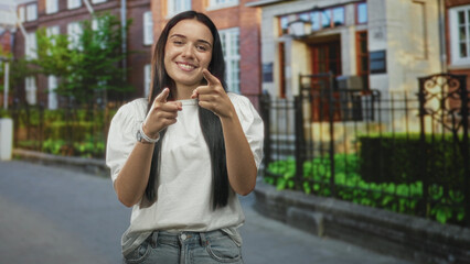 Fototapeta premium Young hispanic woman points index fingers at camera on a residential street by a building and iron fence, wearing a white tshirt and bracelet; confidence joy.