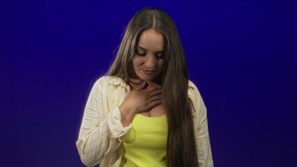 Fototapeta premium Woman with hand on chest and long hair wearing yellow tank top and open striped shirt, soft smile in blue studio; gratitude.