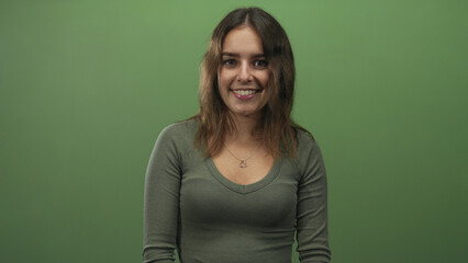 Fototapeta premium Woman smiling, young, wearing heart pendant necklace and olive green top against green studio wall; cheerful warmth.