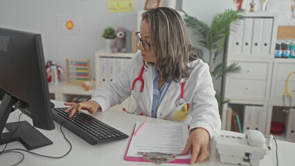 Hispanic woman doctor with grey hair and glasses typing on keyboard with fingers, organizing clipboard and papers at desk in medical building; focused care.