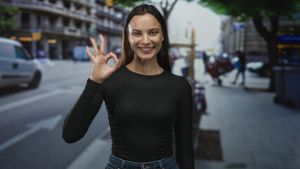 Woman standing facing camera with eyes closed and right hand making ok sign on busy city street, wearing black top and jeans; confidence.
