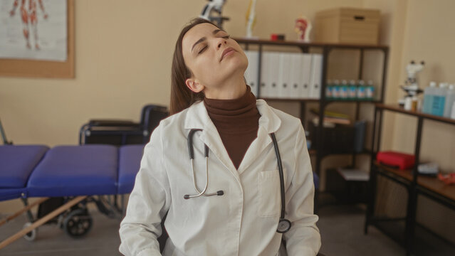 Woman doctor with stethoscope tilting head back showing neck stretch and closed eyes in clinic; clinical fatigue compassion.