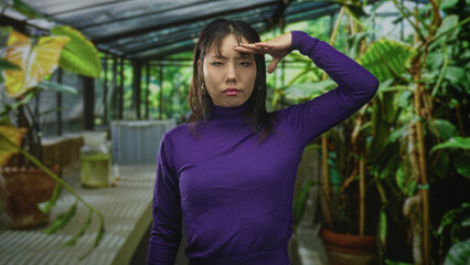 Woman salutes with hand to forehead amid potted plants in a greenhouse building, wearing purple...