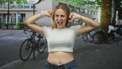 Woman presses fingers into ears with mouth open, bare midriff and navel visible, standing near...