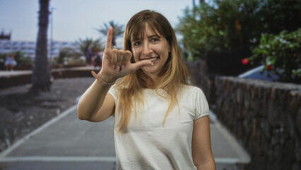 Fototapeta premium Woman showing shaka call me hand sign with visible fingers and wristwatch while smiling on a city street with palm trees and stone wall; playful.