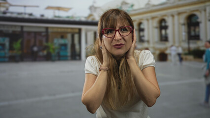 Fototapeta premium Woman covering ears with hands and grimacing in front of ornate building on busy street; frustration travel.