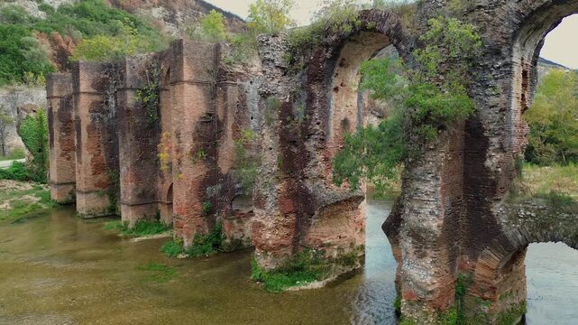 Cinematic drone pullback descending towards the river water, revealing the ancient arches of the Roman Aqueduct of Nicopolis in Epirus Greece, featuring scenic views and historic masonry.