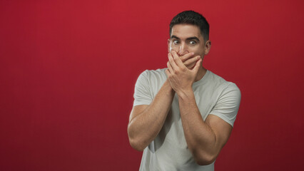 Young hispanic man in gray tshirt with hands covering mouth in red studio; surprise secrecy hush...