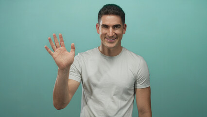 Naklejka premium Man smiling and waving hand to camera in teal studio wearing casual white t shirt with direct eye contact; friendly greeting.