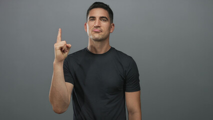 Fototapeta premium Man wearing black tshirt pointing index finger upward in gray studio; determination resolve focus intent.