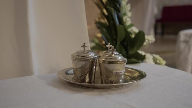 Close view of metal chrism oil containers with small cross lids placed on silver tray over white altar cloth inside catholic church