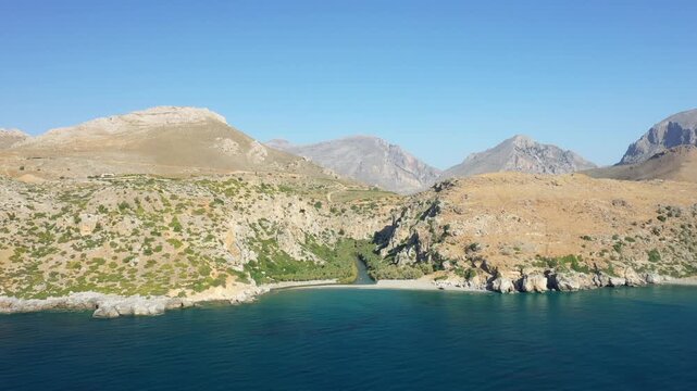 Aerial view of Preveli Beach with rugged mountains, rocky cliffs, and clear blue sea on the south coast of Crete, Greece. The landscape features a river flowing into the Mediterranean surrounded by