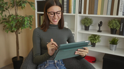 Young woman using tablet with stylus, smiling and tapping screen in a studio with bookshelves and potted plants  learning creativity contentment. © Krakenimages.com