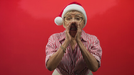 Fototapeta premium Woman in santa hat and glasses cupping hands around mouth while calling out in a studio with vivid red backdrop and striped shirt; holiday joy.