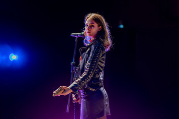 Fototapeta premium A brunette girl in a black leather jacket stands with an electric guitar in front of a microphone, illuminated by stage spotlights against a dark black background, looking directly at the camera.