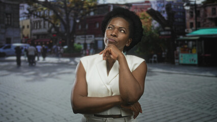 Fototapeta premium Woman with hand on chin and crossed arms in a thinking pose on a city street plaza wearing white sleeveless vest; thoughtful reflection.
