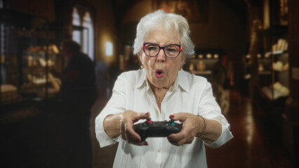 Senior woman holding a gamepad with both hands, thumbs on buttons, focused expression in a museum exhibit hall  playful engagement. © Krakenimages.com