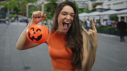 Fototapeta premium Woman holds pumpkin bucket with left hand and makes rock horns gesture with right hand in street; rock fun.