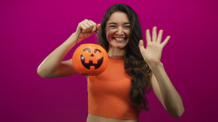 Fototapeta premium Woman in orange top holds jackolantern bucket by hand and waves with smiling grin in studio; happiness.