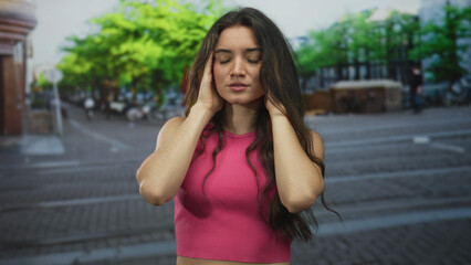 Fototapeta premium Hispanic teenage woman cups cheeks with closed eyes on sunlit urban street with bicycles and trees; pain.