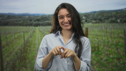 Fototapeta premium Young hispanic woman smiling in a green countryside field forms heart shape with fingers toward camera; love.