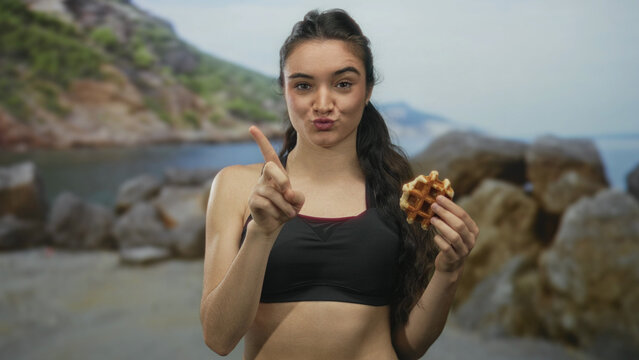 Young hispanic woman holding waffle points finger upward and puckering lips on sunny beach; rejection.