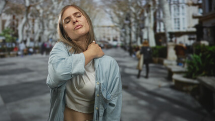 Fototapeta premium Woman holding neck in pain on busy city street with blurred pedestrians, benches, and bare trees; discomfort.