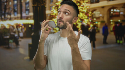 Fototapeta premium Young hispanic man talking on smartphone in urban city street during festive holiday evening with colorful lights and blurred people in the background