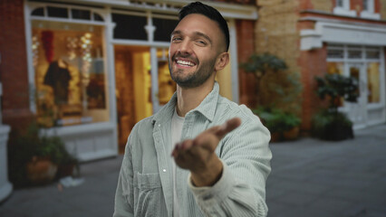 Obraz premium Young man smiling on urban street outdoors invitingly with hand extended wearing casual shirt near shopfront at night capturing warm city ambiance for lifestyle imagery