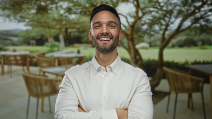Young man with crossed arms and a bright smile stands on a terrace of an outdoor restaurant, surrounded by tables and lush landscape, embodying confidence and leisure. © Krakenimages.com