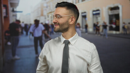Naklejka premium Hispanic man outdoors points with a confident smile in an urban city street setting, wearing a white shirt and glasses, showcasing modern urban living.