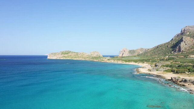 Aerial view of the clear turquoise waters and rugged coastline of Falasarna Beach on the island of Crete, Greece, under a clear blue sky.