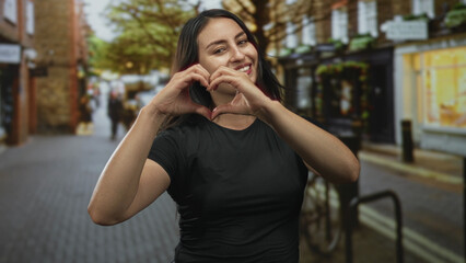 Fototapeta premium Young hispanic woman makes heart with hands on a busy city street near a bicycle rack while smiling broadly; love joy.