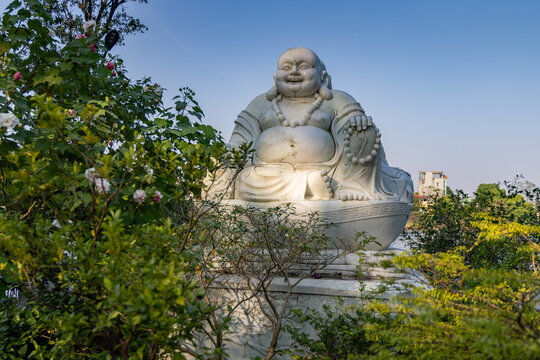 A large white statue of the smiling Laughing Buddha, symbolizing happiness and prosperity, stands amidst green foliage at Non Nuoc Pagoda,  Ninh Binh, Vietnam.