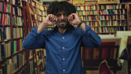 Man with curly hair rubbing his eyes among tall bookshelves in a library building, wearing a blue shirt and showing a weary grimace  study fatigue. © Krakenimages.com